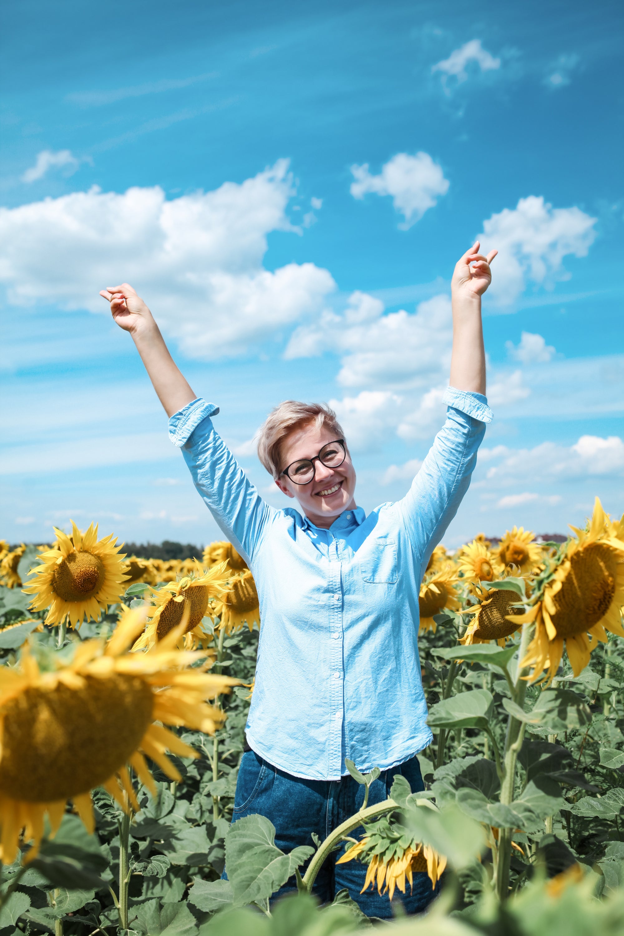 Person with arms raised in a sunflower field under a blue sky with clouds